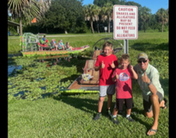 Captain Mike posing with two children with an airboat them on the water   